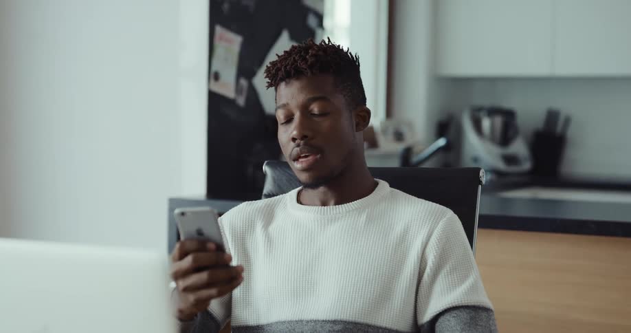 A man is sitting at a table in front of a laptop computer and he answers his cell phone