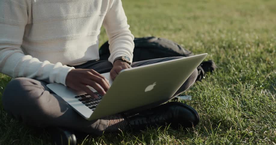 A man is sitting in a field and typing on laptop