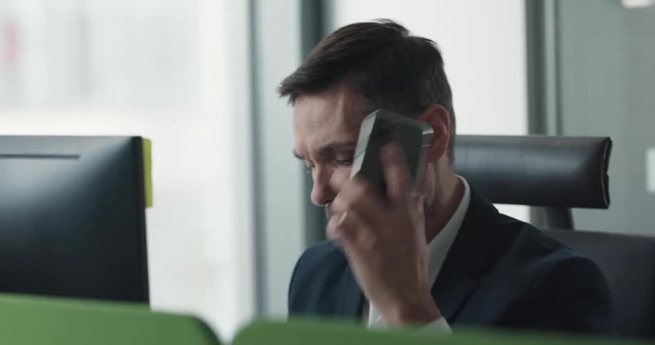 Male office worker sitting at his desk next to the window working on his computer handling some papers and talking on the phone