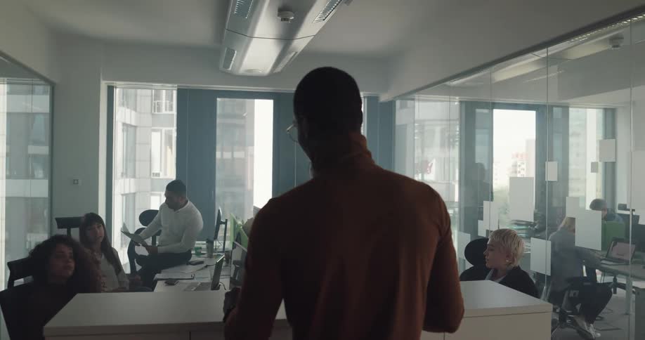 A man holding a stack of papers speaks to an office of people sitting at their desks during the day