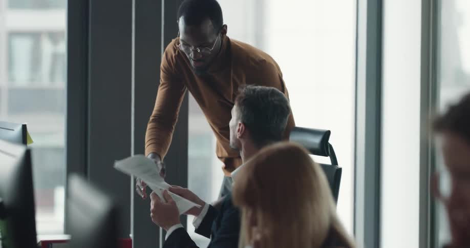 A man walks over to another man in an office and gives him paperwork