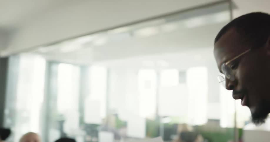 A man standing in an open office space talking and gesturing with paper as he talks to the team