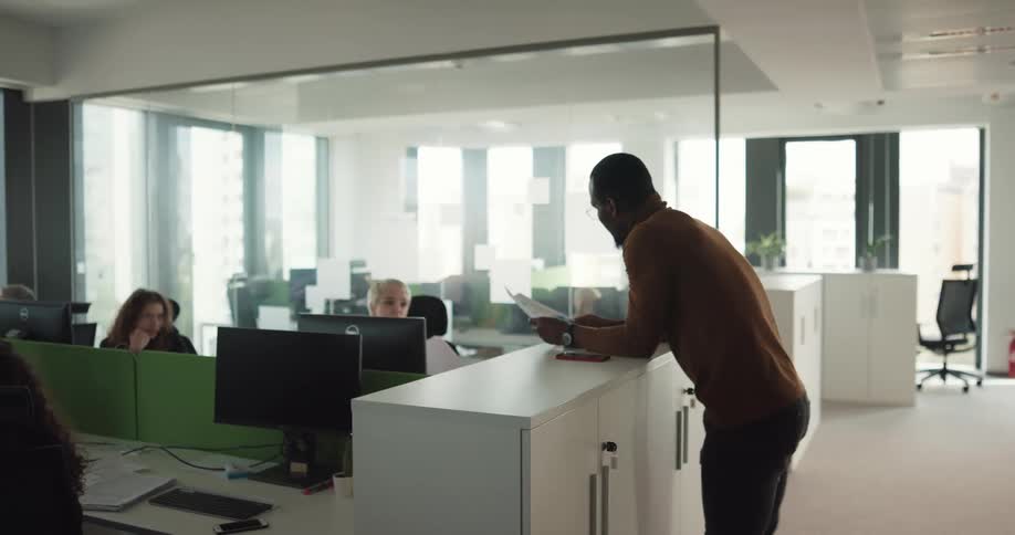 A bunch of women are sitting at desk infront of computers when a man starts passing out papers