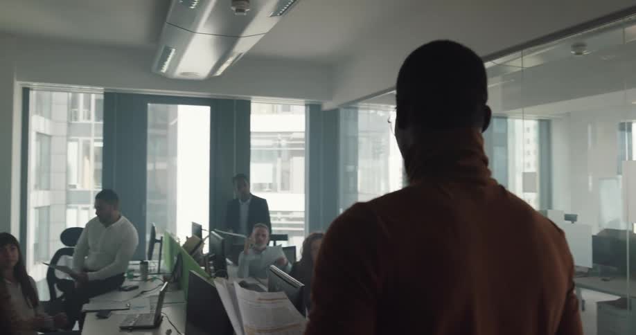 Black man holding some papers while standing in front of a meeting table talking with his coworkers in an office meeting