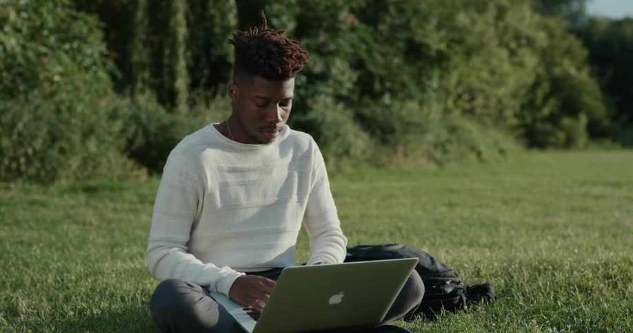 A man is sitting in the grass and typing on his laptop
