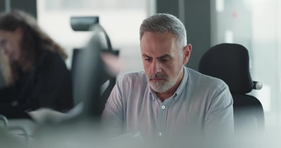 A man sitting in an office at a desk near a woman is upset as he looks at paperwork