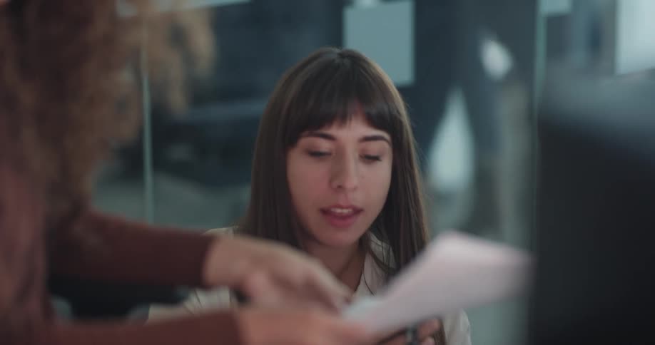Two female coworkers sitting in an office space having a conversation and handling some papers while one of them is using her smartphone