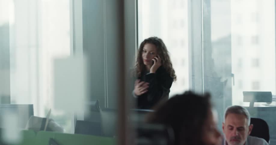 A woman standing in an office raises her hand to her face in distress while talking on her phone
