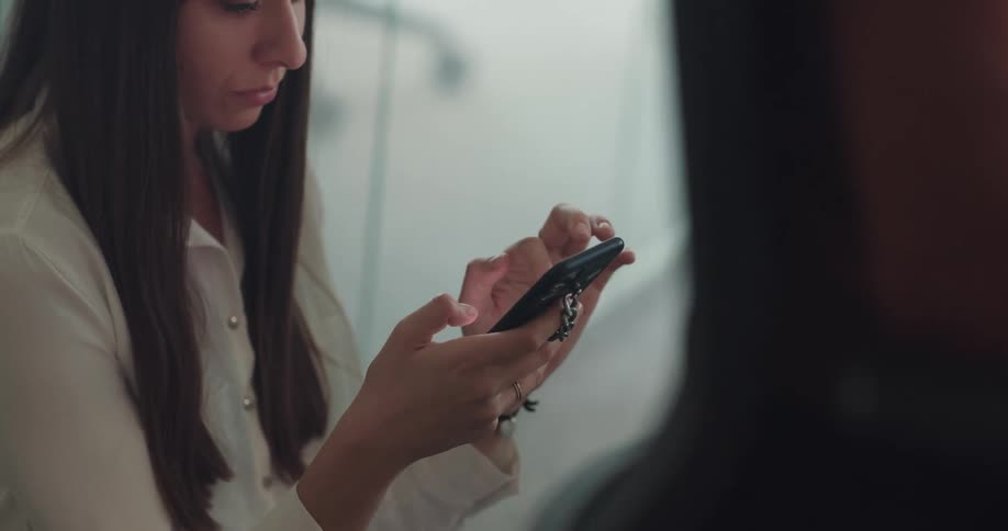 Young female office worker sitting near a glass door in the office using her smartphone