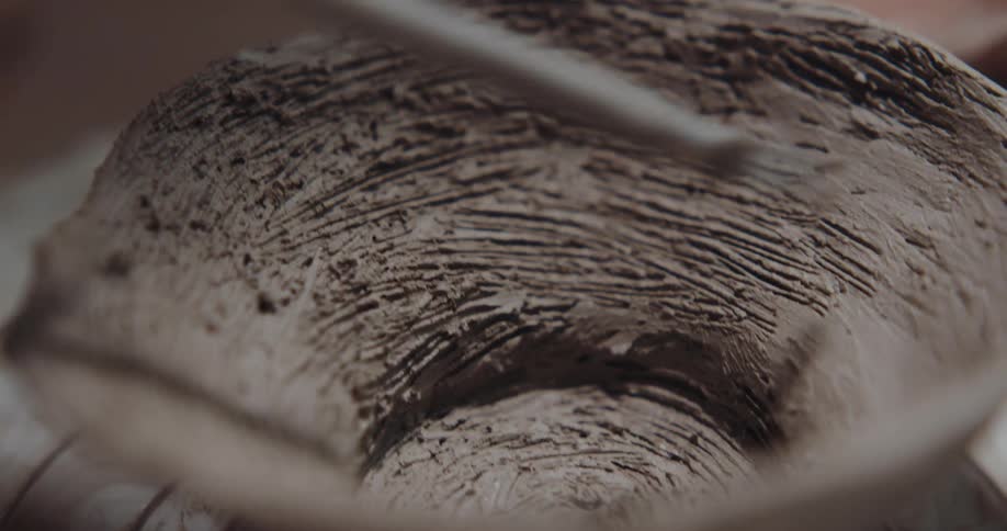 A person turns a clay bowl on a wheel as they use a tool to shape it