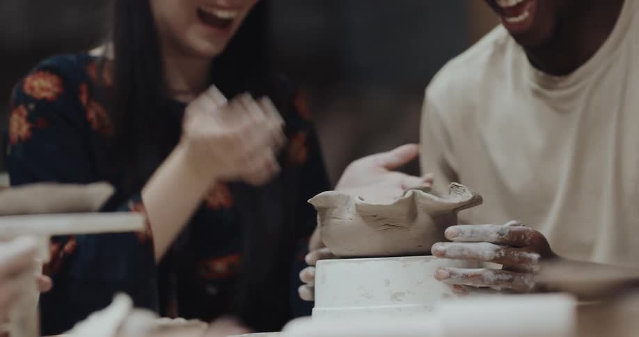 A man and woman laugh as they work on creating a clay bowl