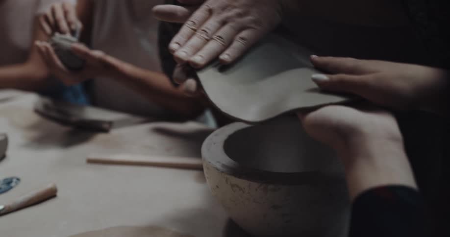 Two people pick up a soft clay disk to place it over a ceramic bowl