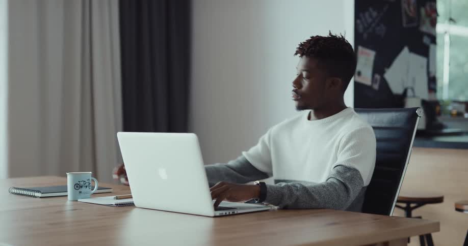 A man is sitting at a table in front of a laptop computer and he is writing on paper and he drinks his coffee