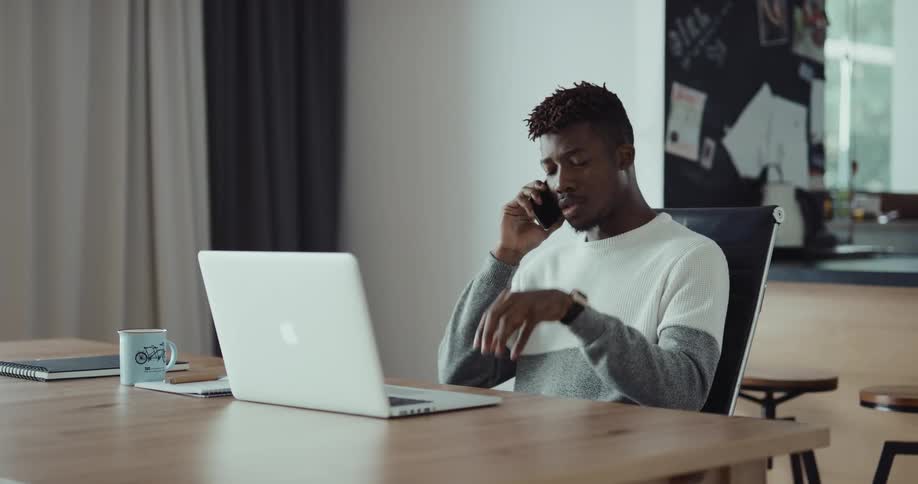A man is sitting at a table in front of a laptop computer and he answers his cell phone