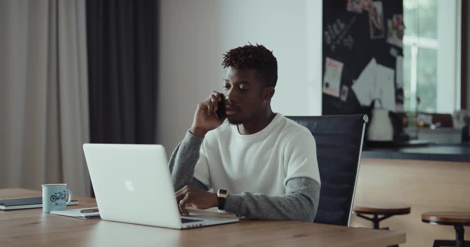 A man sits at a large desk with a laptop and speaking on the phone