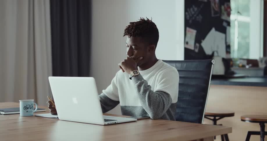 A man is sitting at a table in front of a laptop computer and he is writing on paper