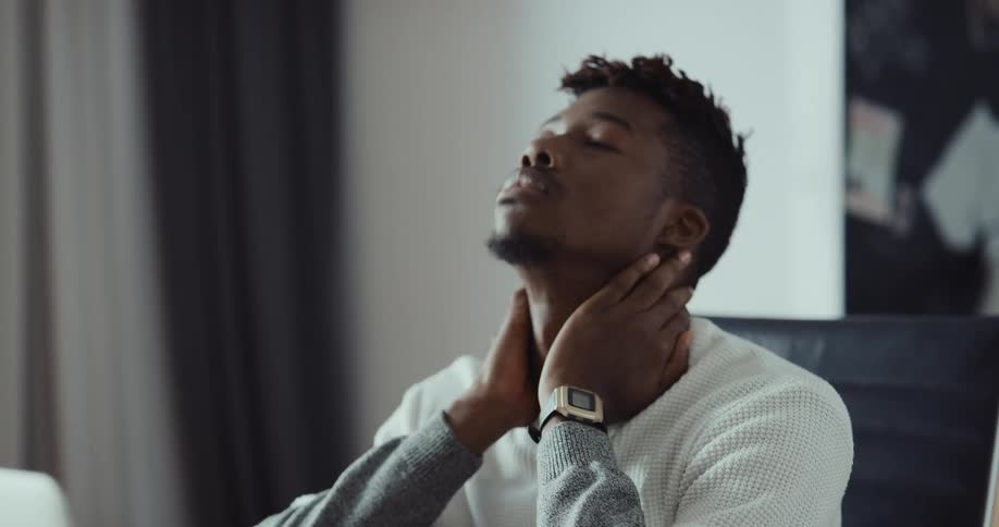Man with white and gray shirt types on laptop and rubs his neck sitting at table