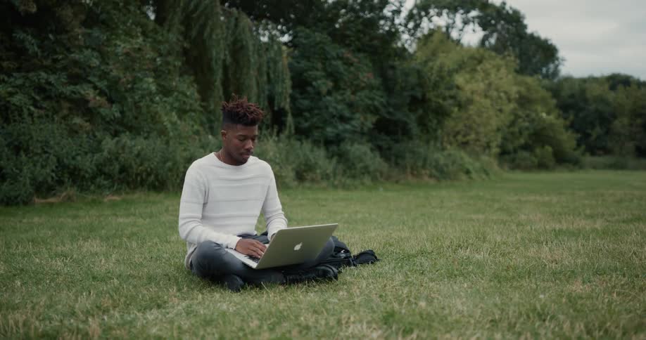 A man is typing on a laptop and sitting in a field