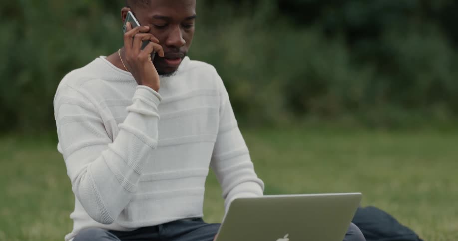 A man sits in a grassy area with a laptop and picks up a cell phone to talk