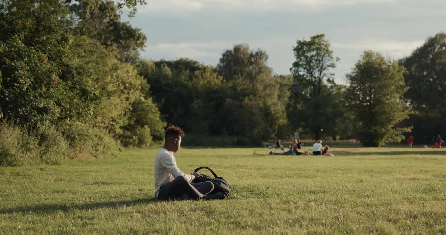 A man sits down in the grass and uses his laptop