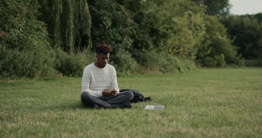 A man is sitting outside on the grass with his laptop computer and he sets it on the grass and picks up his cell phone
