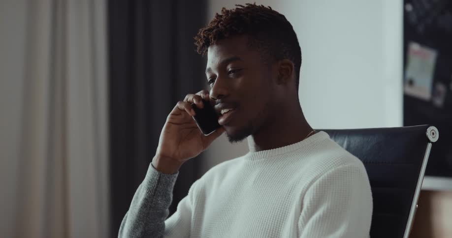 A man is sitting at a table in front of a laptop computer and he answers his cell phone