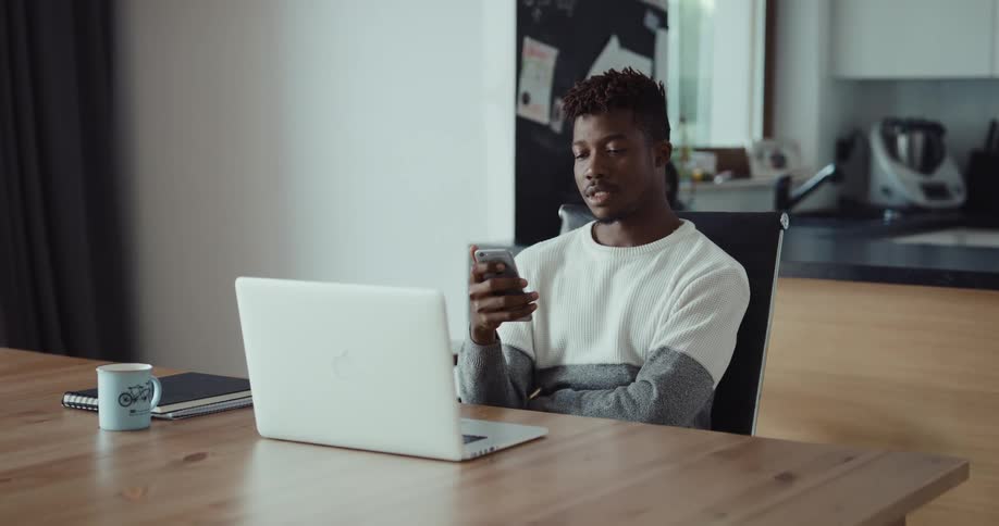 Black man sitting at the desk with a laptop in front of him talking on his smartphone