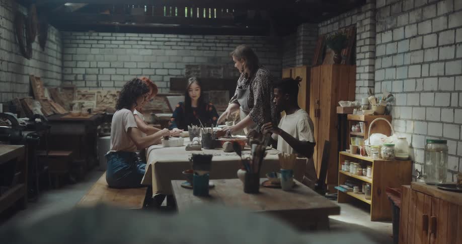 A group of people is sitting at a table preparing food while a woman walks around helping them