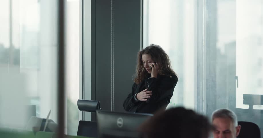 Young female office worker standing near the glass window in the office talking on the phone