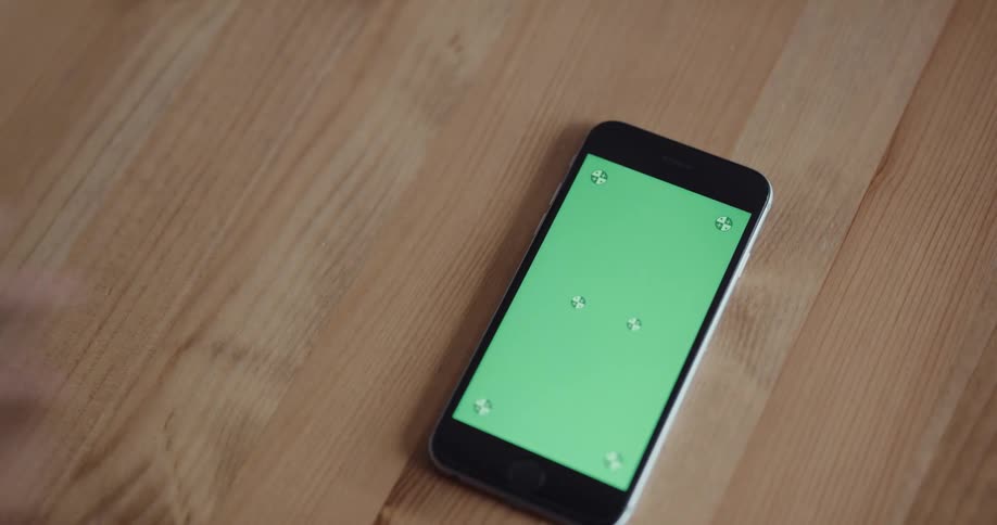 A cell phone with a green screen is sitting on a wooden table and someone picks it up and sets it back down