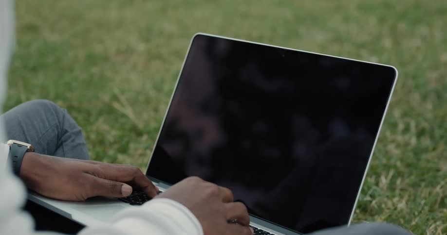 A man is sitting outside on the grass typing on his laptop computer