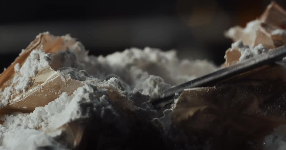 A person picks up white cocaine powder from an open bag with a knife