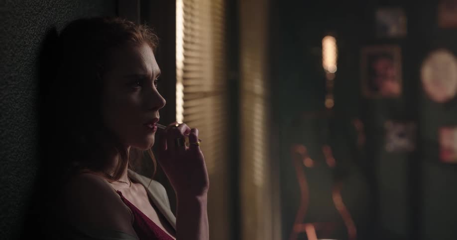 Young woman leaning against the wall next to a window with a thoughtful facial expression lighting up a cigarette and starting to smoke