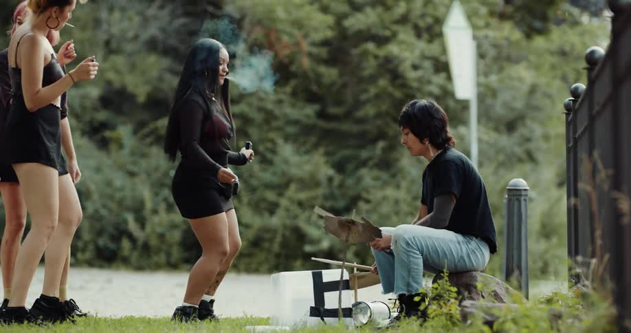 Group of female friends and a young man dancing and partying in a public park while the man is sitting on a rock and playing a musical instrument at a sunny day