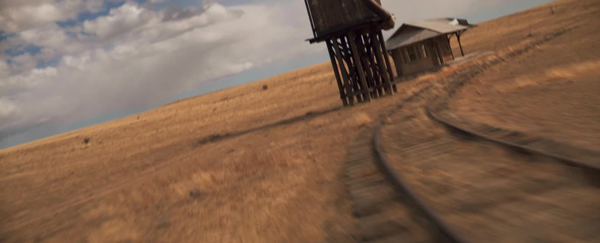 Train track curves past station and wooden water tower in field with mountains in the background