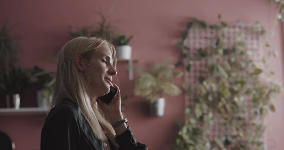 Happy young blonde woman talking on the phone while standing next to a wall full of potted plants