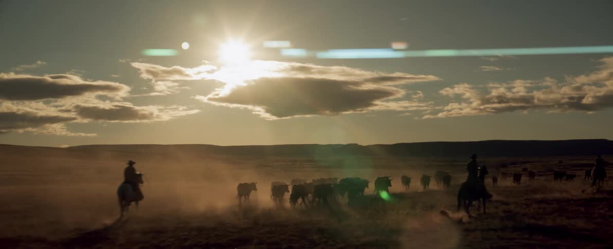 Three cowboys drive herd of cattle across plains as sun shines from cloudy sky