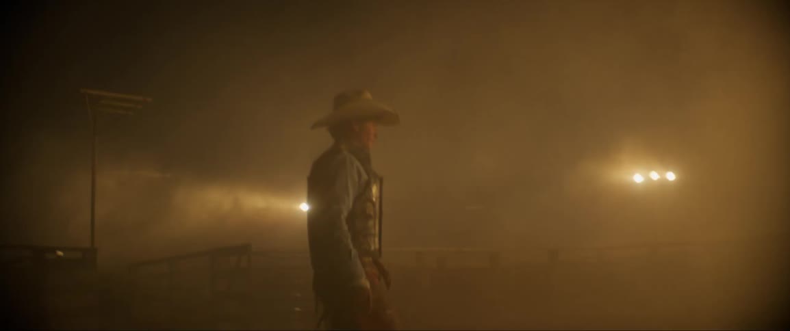 A man wearing a badge walks towards sets of lights in the dust