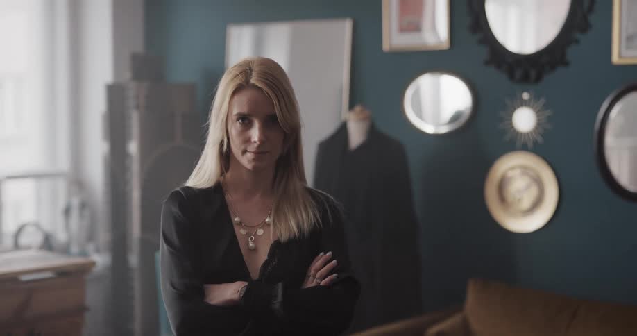 A woman standing indoors looks into the camera for a moment before crossing her arms and smirking