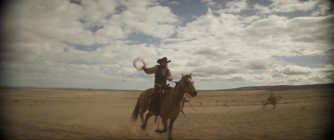 A trio of cowboys ride horses through a grassland during the day