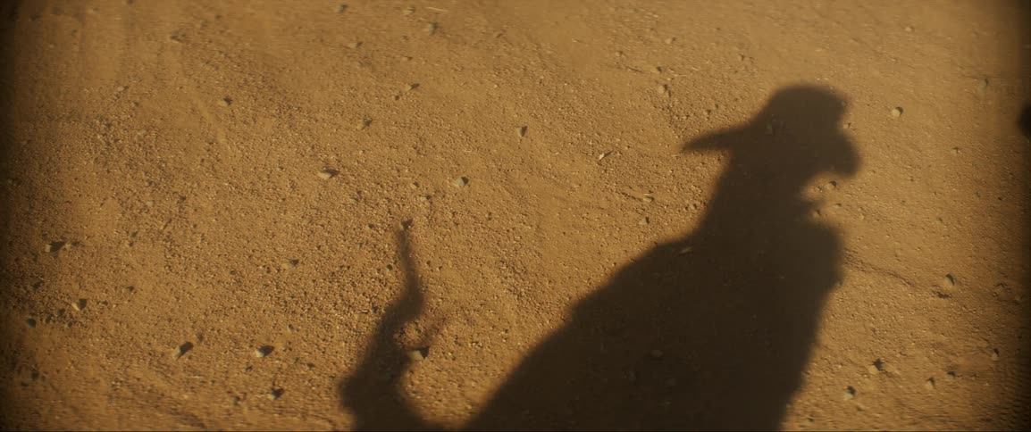A shadow of a man on sand as he shoots a gun several times and then falls backwards as he is hit