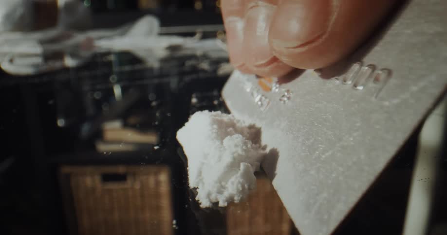 A right hand scrapes a powdery white substance nervously on a mirrored table with a credit card eventually creating one line