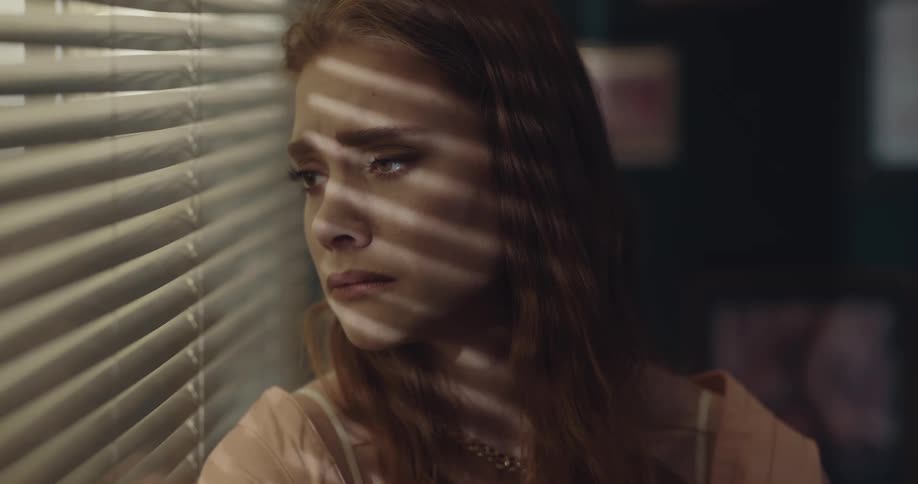A woman looks sad as she looks out a window between the blinds