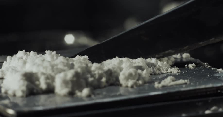 Cocaine is sitting on a mirror as someone scoops up some of the powder with a knife blade