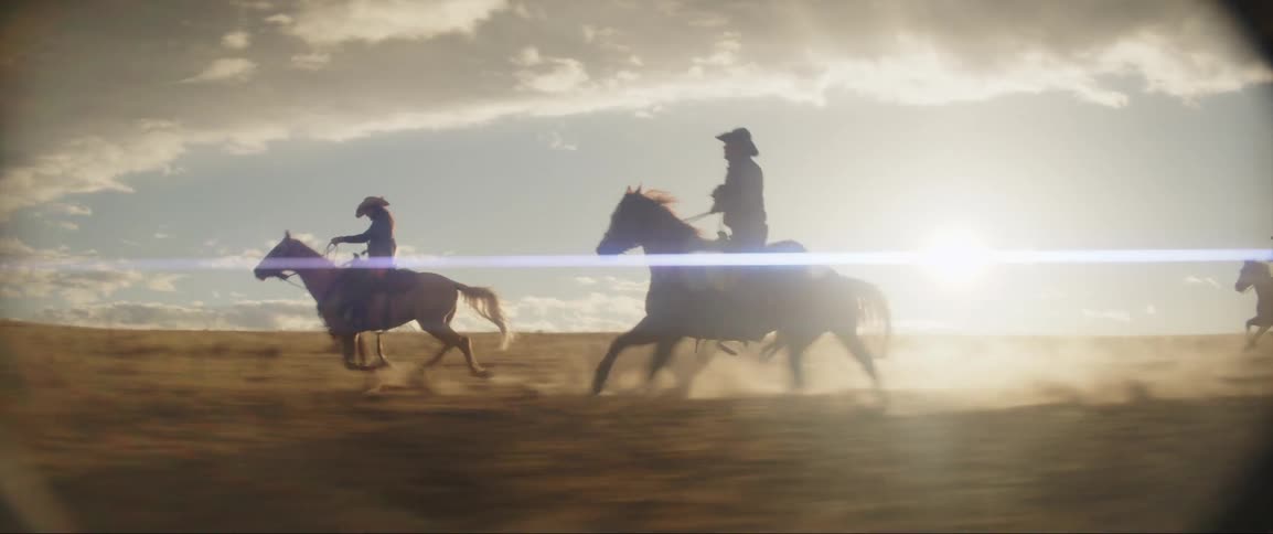 A group of cowboys ride horses across a dusty grassland during the day