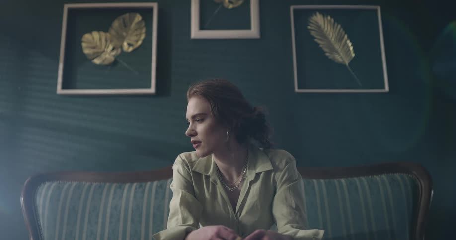 A woman sits on a couch in a dark teal room slightly fidgeting with her hands glancing towards the light and then glancing down