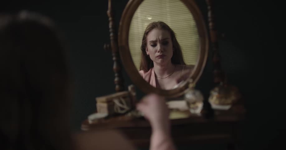 Young redhead woman with a sad expression on her face sitting in front of a round mirror and taking her earrings off