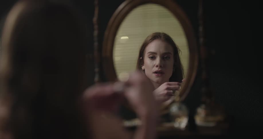 Young redhead woman applying makeup and checking herself on a round mirror on a dressing table