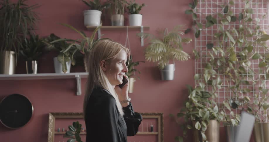 Young blonde woman standing indoors next to a pink wall filled with potted plants talking on the phone