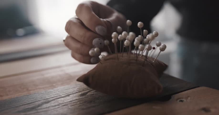 A brown velveteen pincushion filled with pearl pins sits on a table and a standing woman in jeans inserts and removes pins